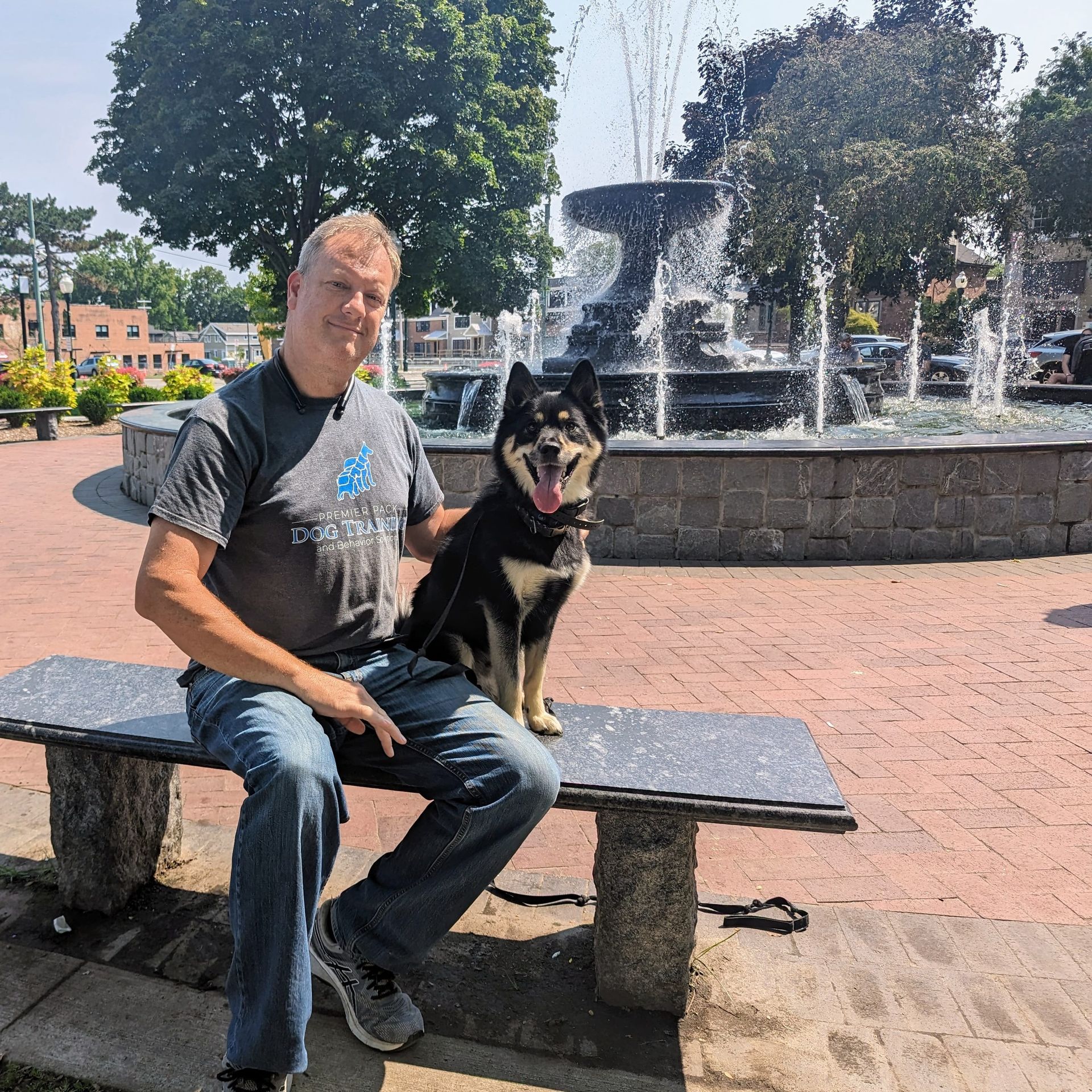 A man is sitting on a bench with a dog in front of a fountain