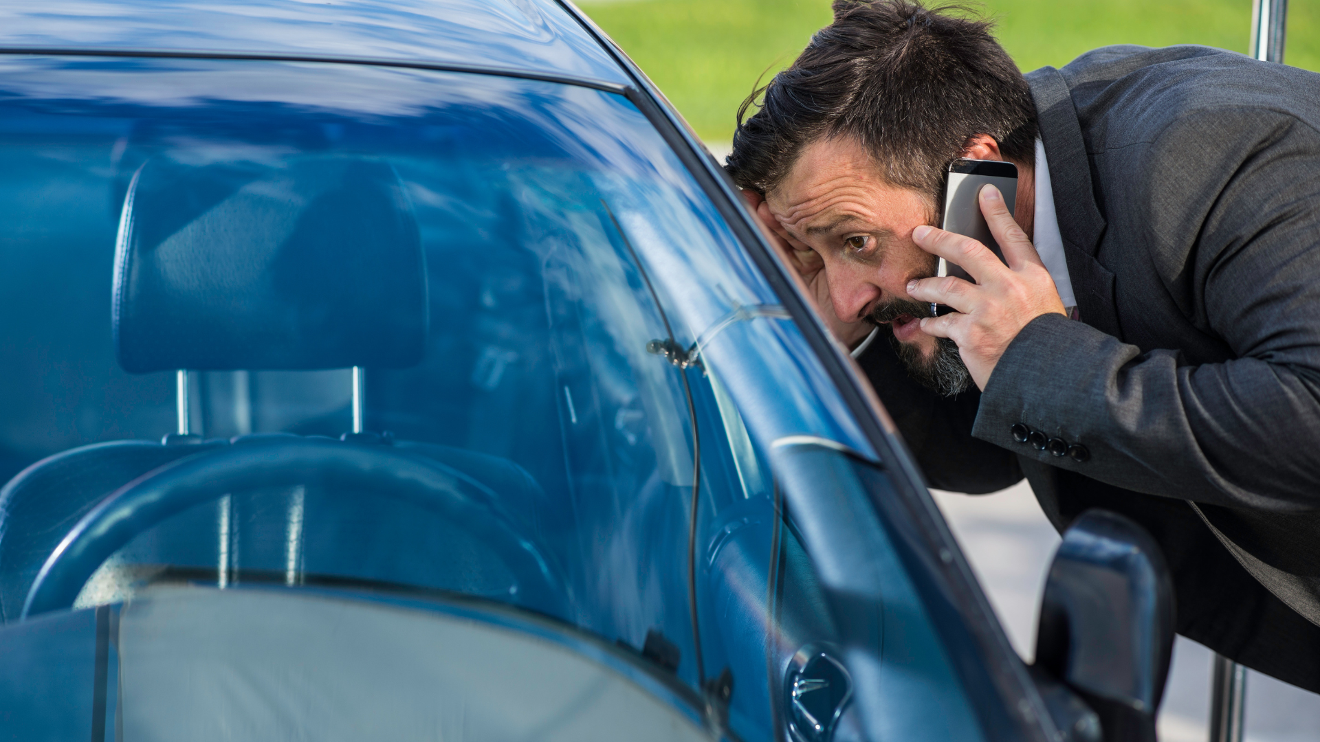 Man in suit, on phone, peers intently into a car, possibly locked, looking for something.