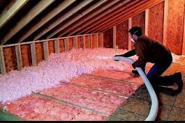 Insulation being blown into an attic from a white hose, covering the floor.