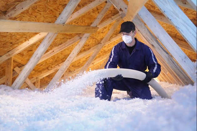 Insulation being blown into an attic from a white hose, covering the floor.