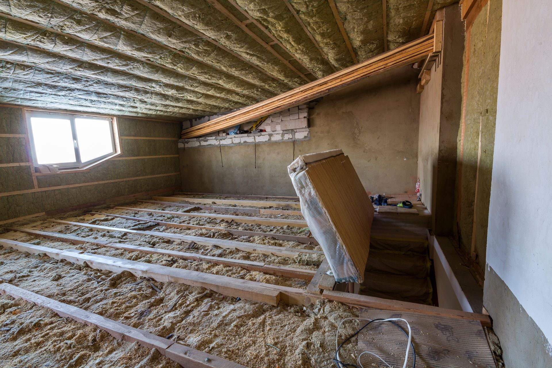 Interior of an unfinished attic with exposed beams, insulation, and a window. Interior of an unfinished attic with exposed beams, insulation, and a window.