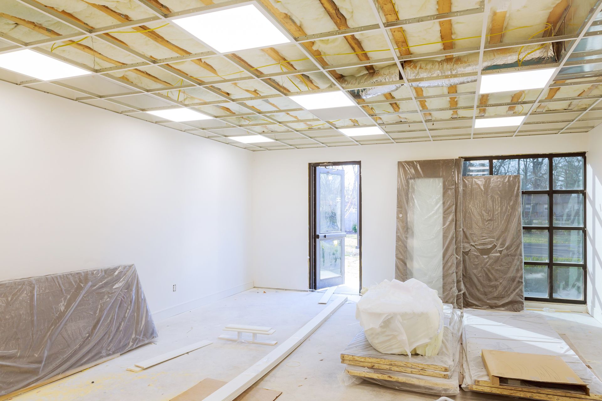Room under construction with exposed ceiling grid, bright lights, white walls, and debris on floor. Room under construction with exposed ceiling grid, bright lights, white walls, and debris on floor.