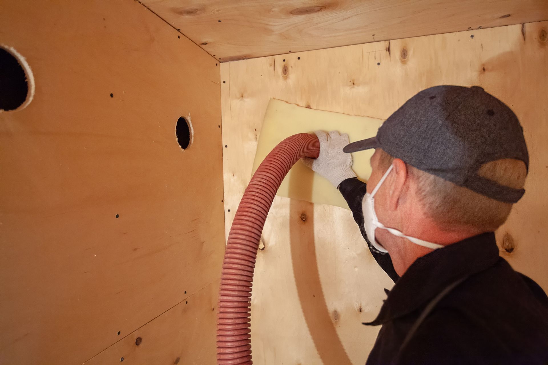 Person installing insulation in a wooden structure, using a hose. They wear a mask, gloves, and a hat. Person installing insulation in a wooden structure, using a hose. They wear a mask, gloves, and a hat.