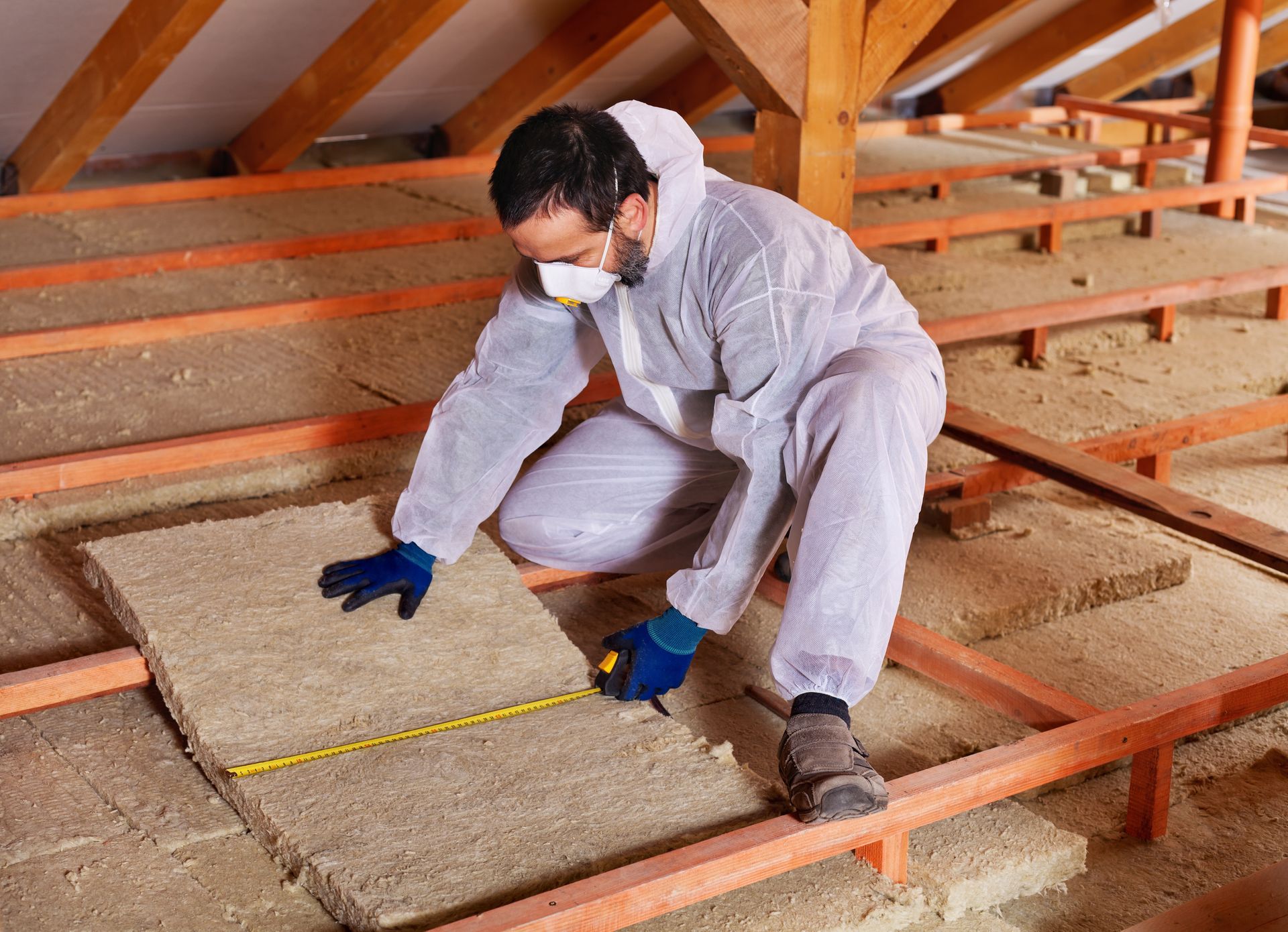 Man in protective suit measuring insulation in an attic, wooden beams visible.