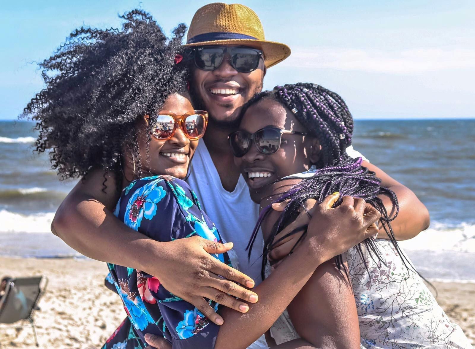 Three people embrace on a sunny beach, smiling. One wears a hat and sunglasses, two wear sunglasses. Ocean in the background.