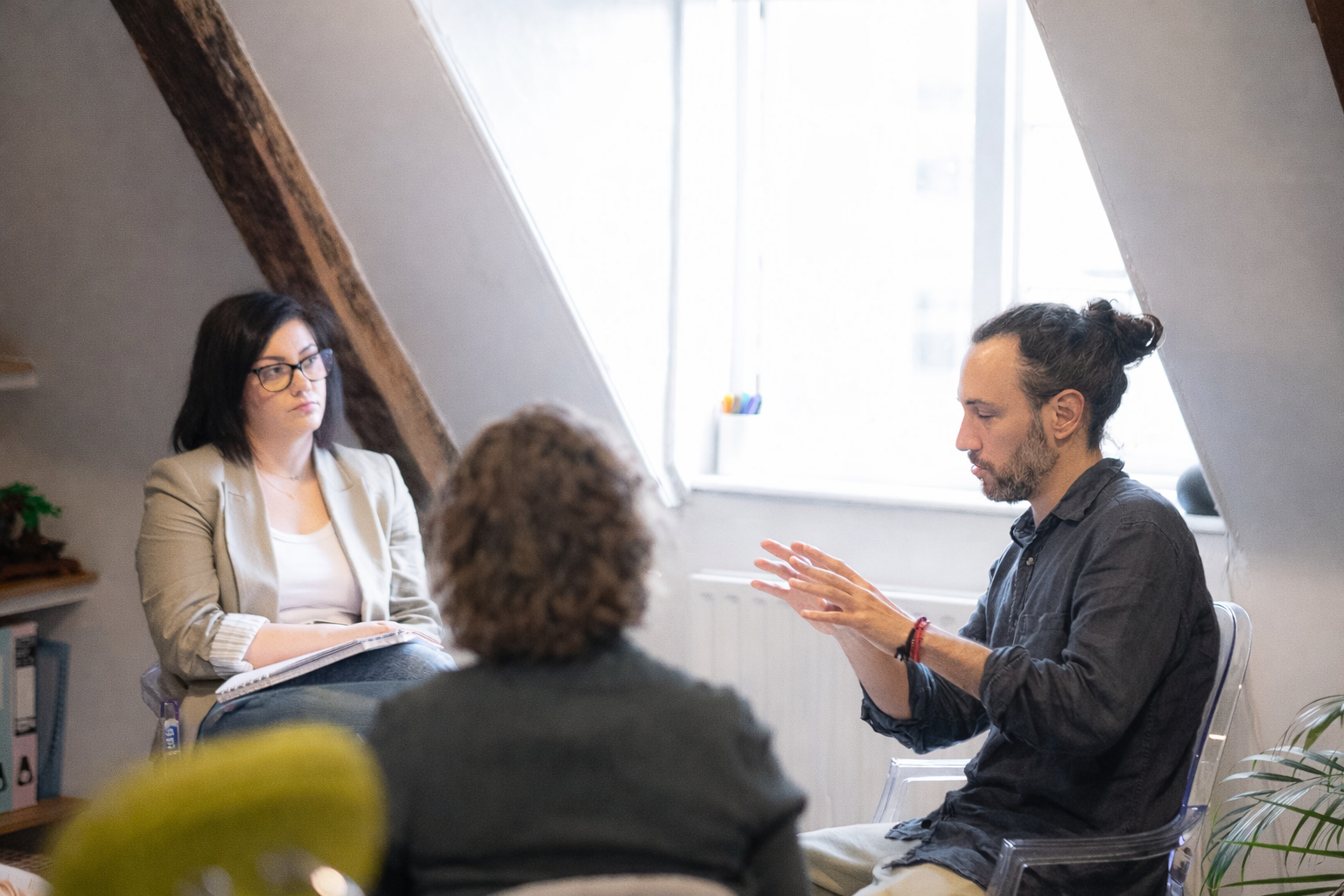 Three people in a room, two seated facing a man gesturing with hands, possibly a discussion or counseling setting.
