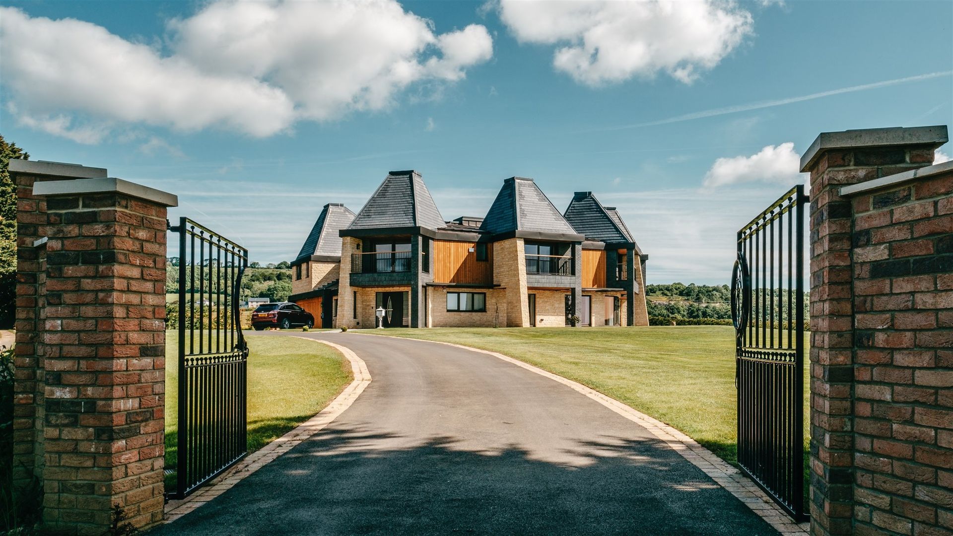 Large modern house with open black gates and a long driveway on a sunny day.