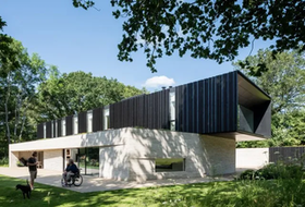 Modern two-story home with cantilevered upper level, dark wood siding, concrete base, and people, including one in a wheelchair, in front.