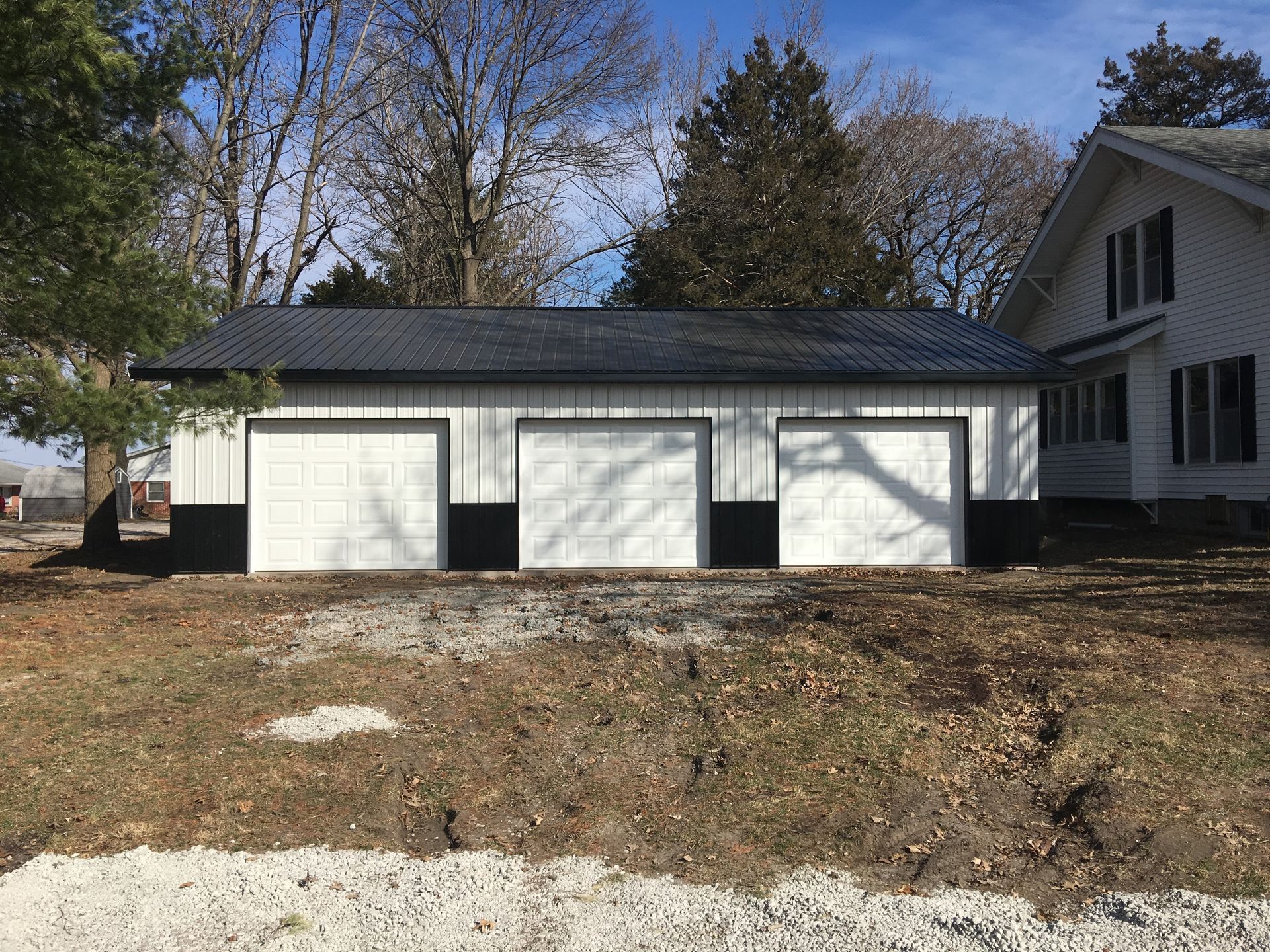 A black and white garage with a black roof is in a dirt field next to a house.