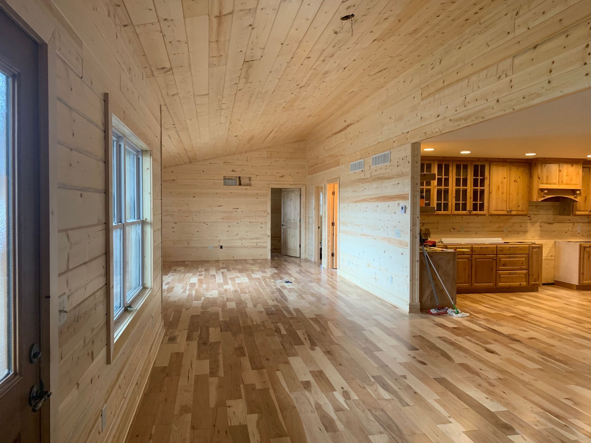 A large empty room with hardwood floors and a kitchen in the background.