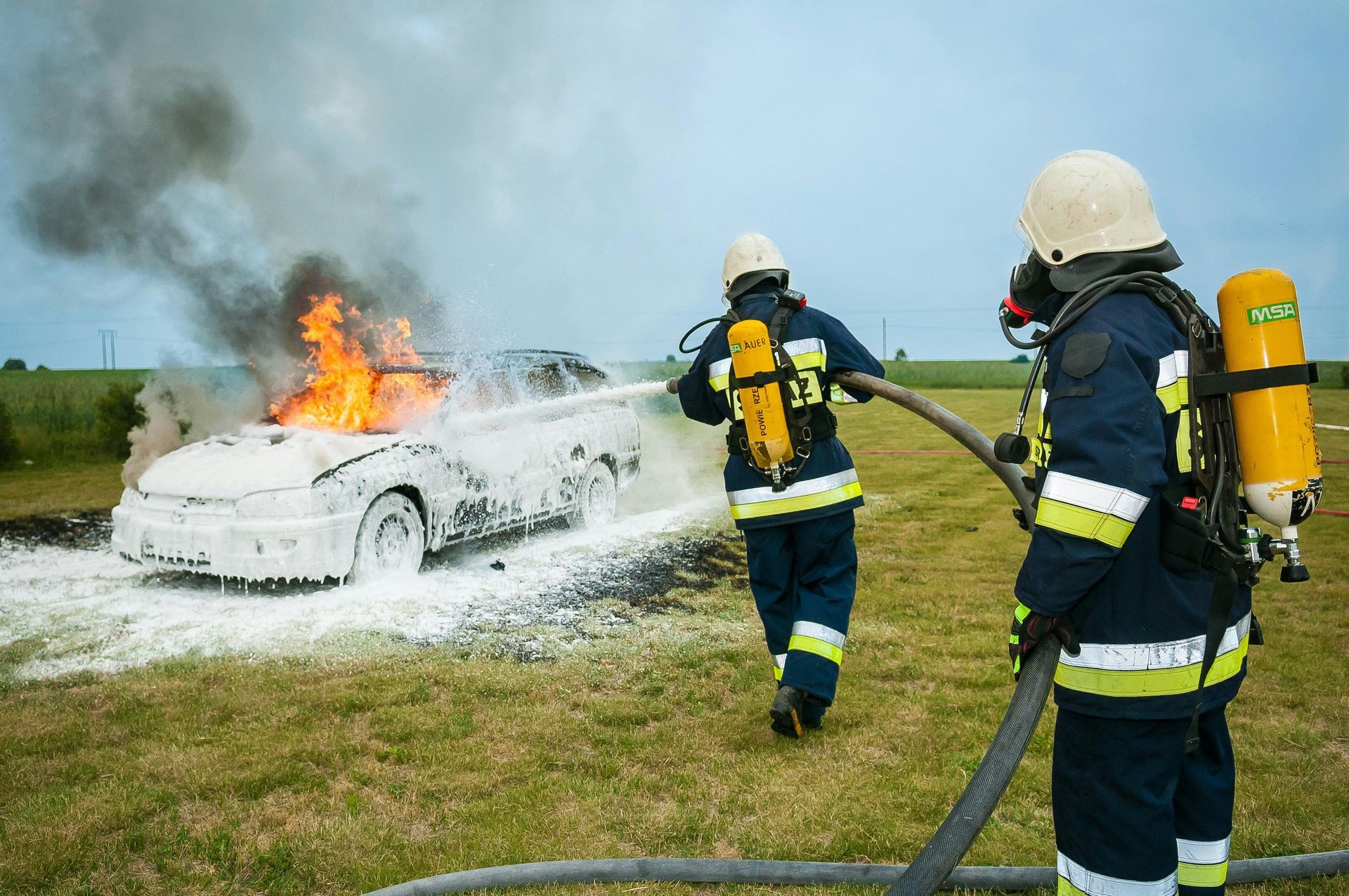 Two firefighters are fighting a car fire with a hose.