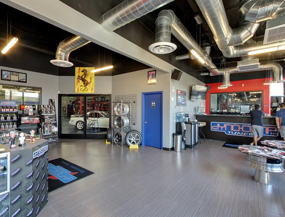 Interior of an auto shop with a vintage car visible through a doorway, and people near a service counter. | Dyno-Comp