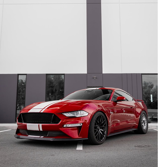 A red Ford Mustang with white racing stripes parked on asphalt in front of a modern building with gray and white walls. | | Dyno-Comp