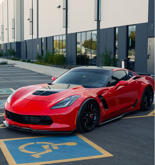 A red Chevrolet Corvette Z06 parked in a handicap-accessible spot in front of a modern commercial building. | | Dyno-Comp