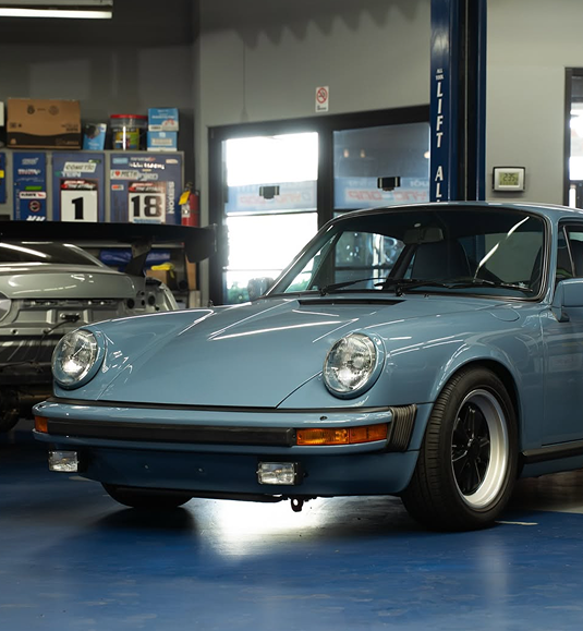 A light blue Porsche 911 parked inside a garage, with a second car and storage cabinets visible in the background. | Dyno-Comp