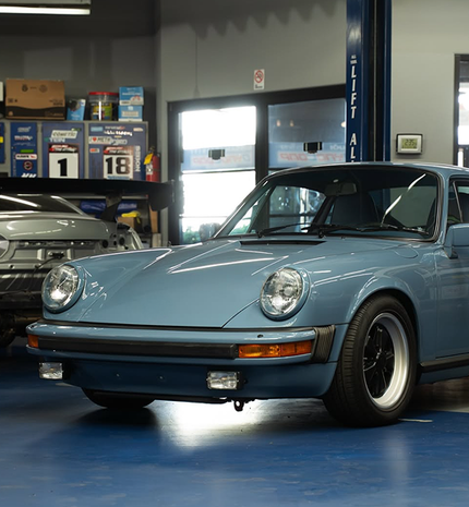 A light blue Porsche 911 parked inside a garage, with a second car and storage cabinets visible in the background. | Dyno-Comp