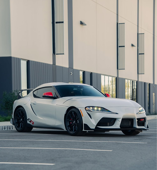 A white Toyota GR Supra with red side mirrors parked in an empty parking lot beside a modern industrial building. | Dyno-Comp