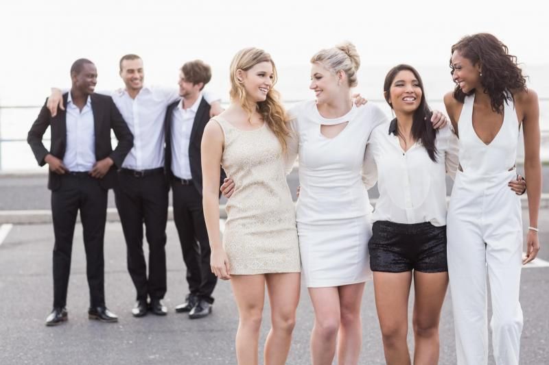 A group of friends laughing and standing together outdoors, with three men in formal wear and four women in light clothing.