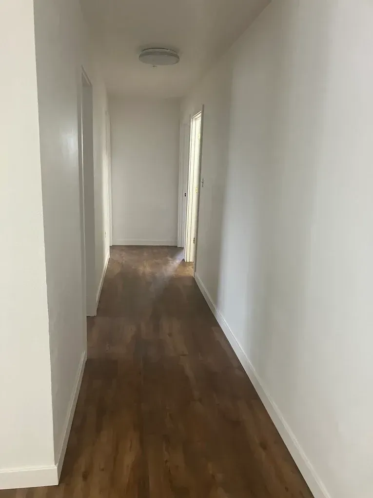 A bright hallway with white walls and wood-patterned flooring, leading toward a doorway with a decorative curtain.