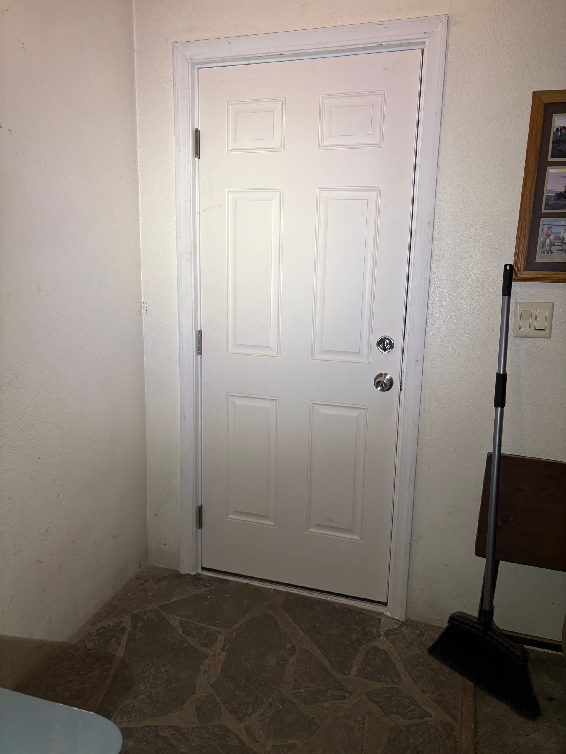 A white six-panel door with silver hardware set in a room with textured walls and a stone tile floor, near a broom.