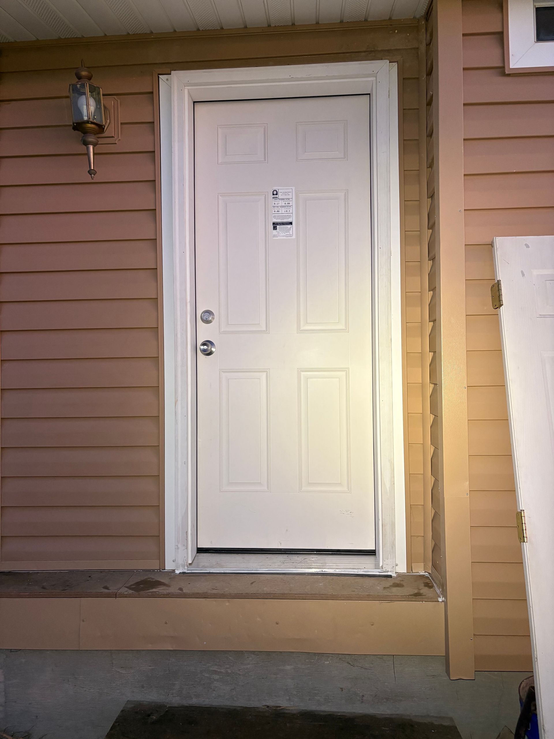 A white entry door with a rectangular window installed in a tan house with horizontal siding and a concrete step.