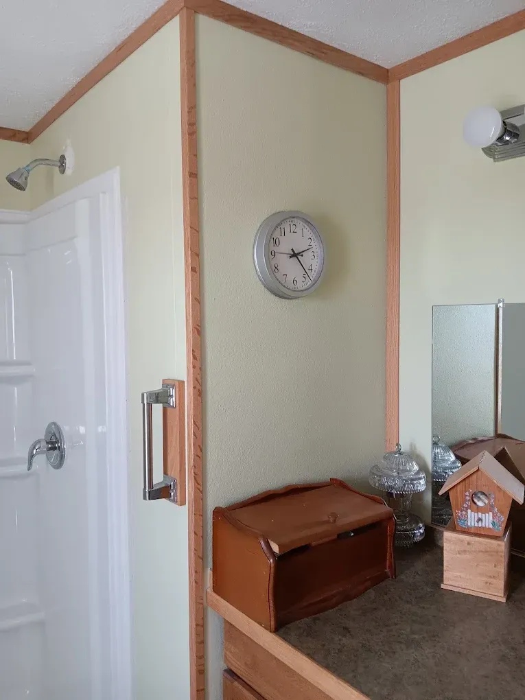 A bathroom corner with light yellow walls, a shower stall, a wall clock, and a wooden bread box on the vanity counter.