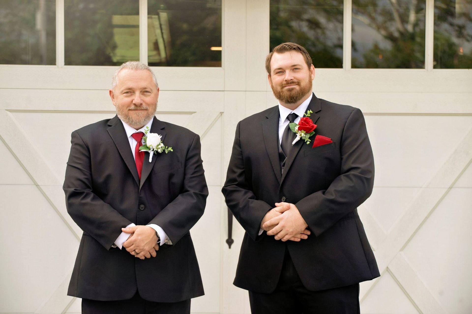 Two men in suits stand in front of a white garage door, both with floral boutonnieres.