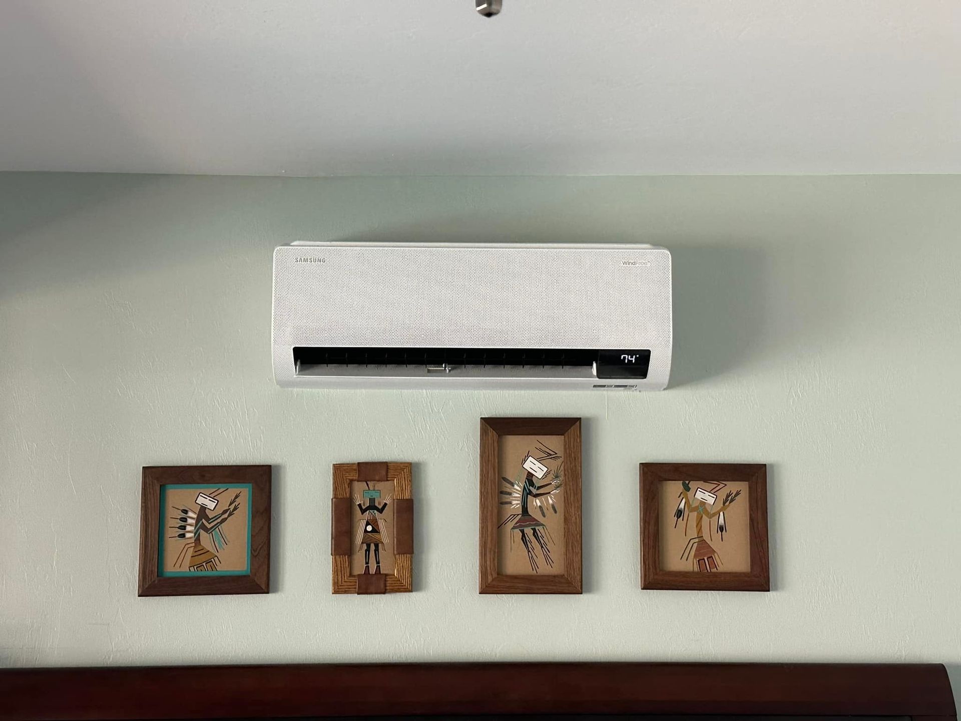 Air conditioner above four framed art pieces on a light green wall.