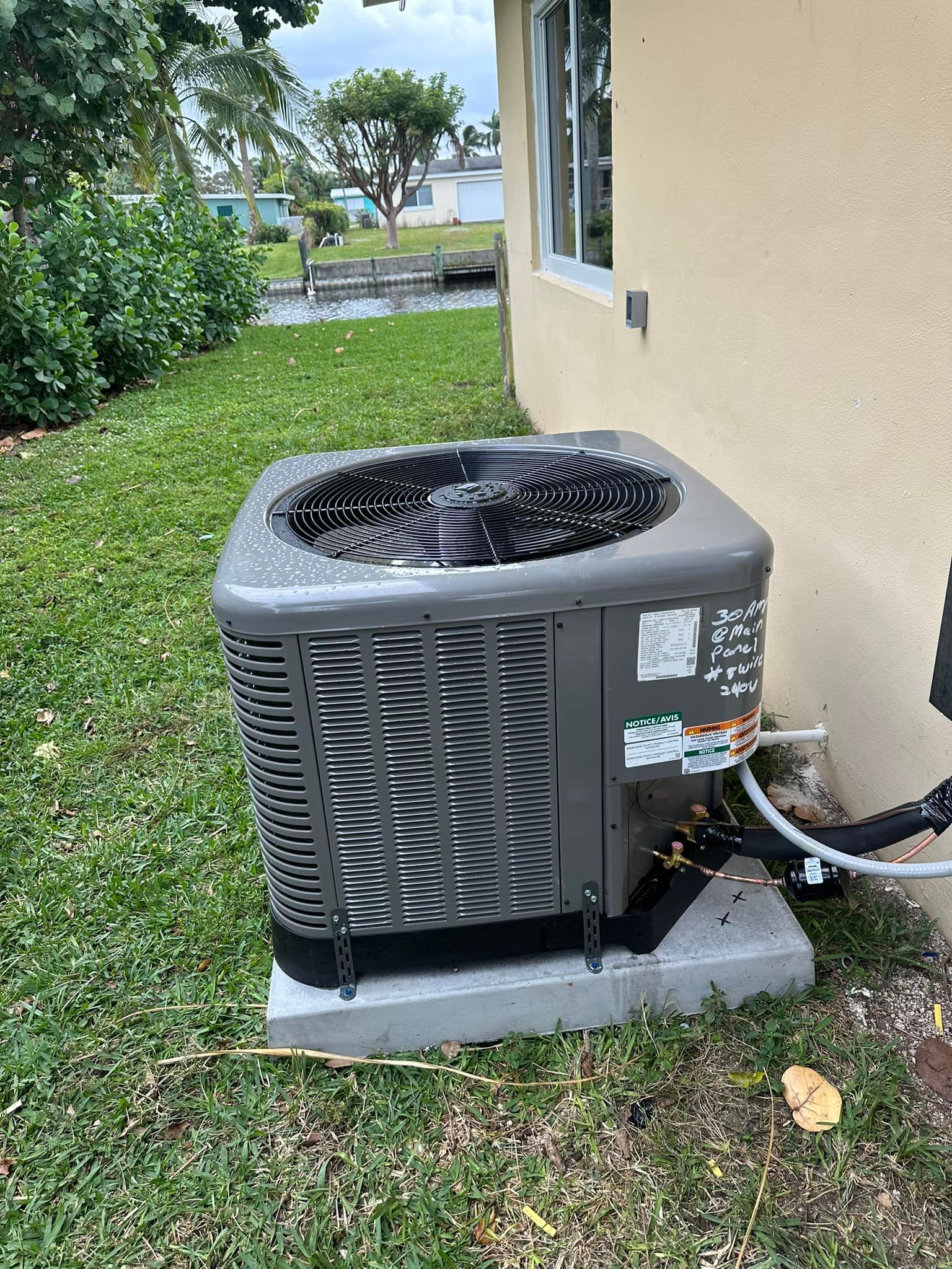 An air conditioning unit on a concrete pad next to a light-colored building, with green grass and a canal in the background.