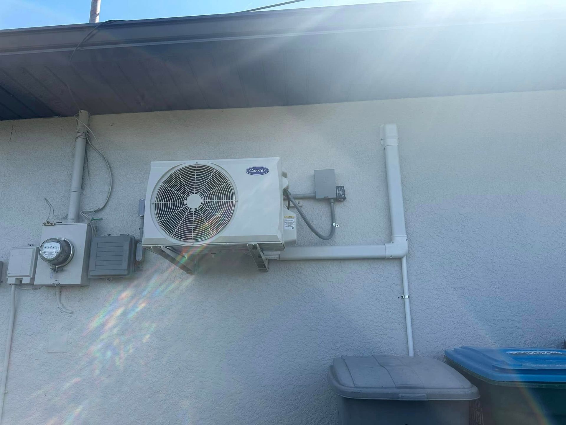 White air conditioner mounted on a stucco wall next to electrical components and a trash bin.