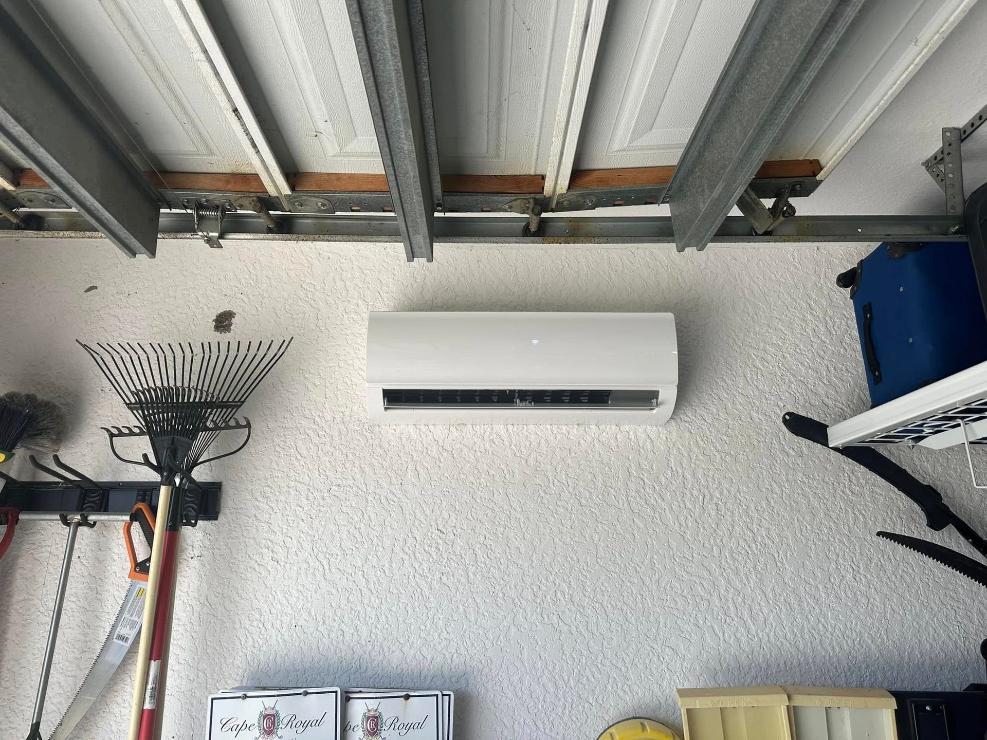 White mini-split air conditioner mounted on textured garage wall with tools and garage door components visible.