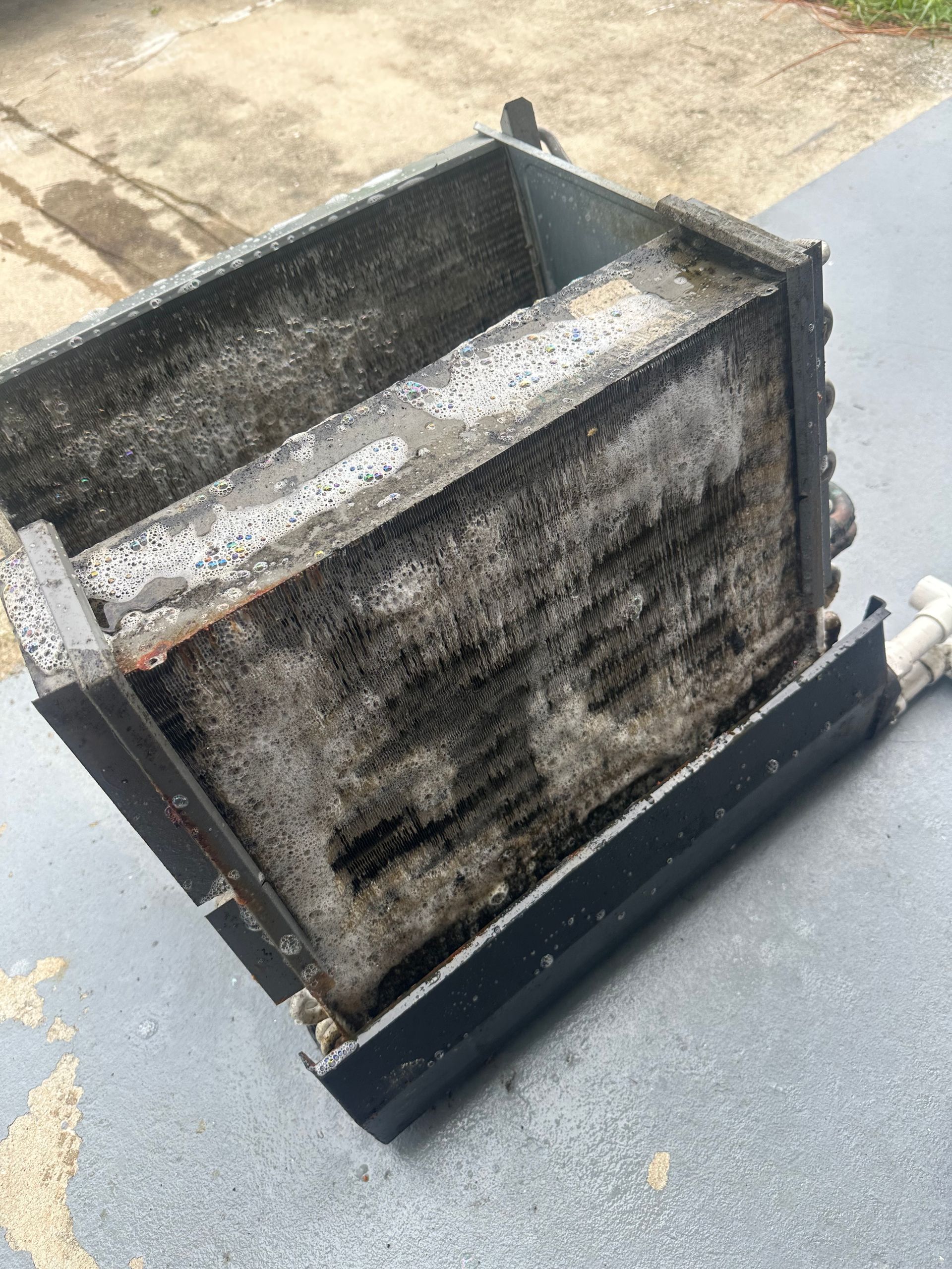 A rectangular, dirty HVAC evaporator coil outside on a surface. It is gray with black markings.