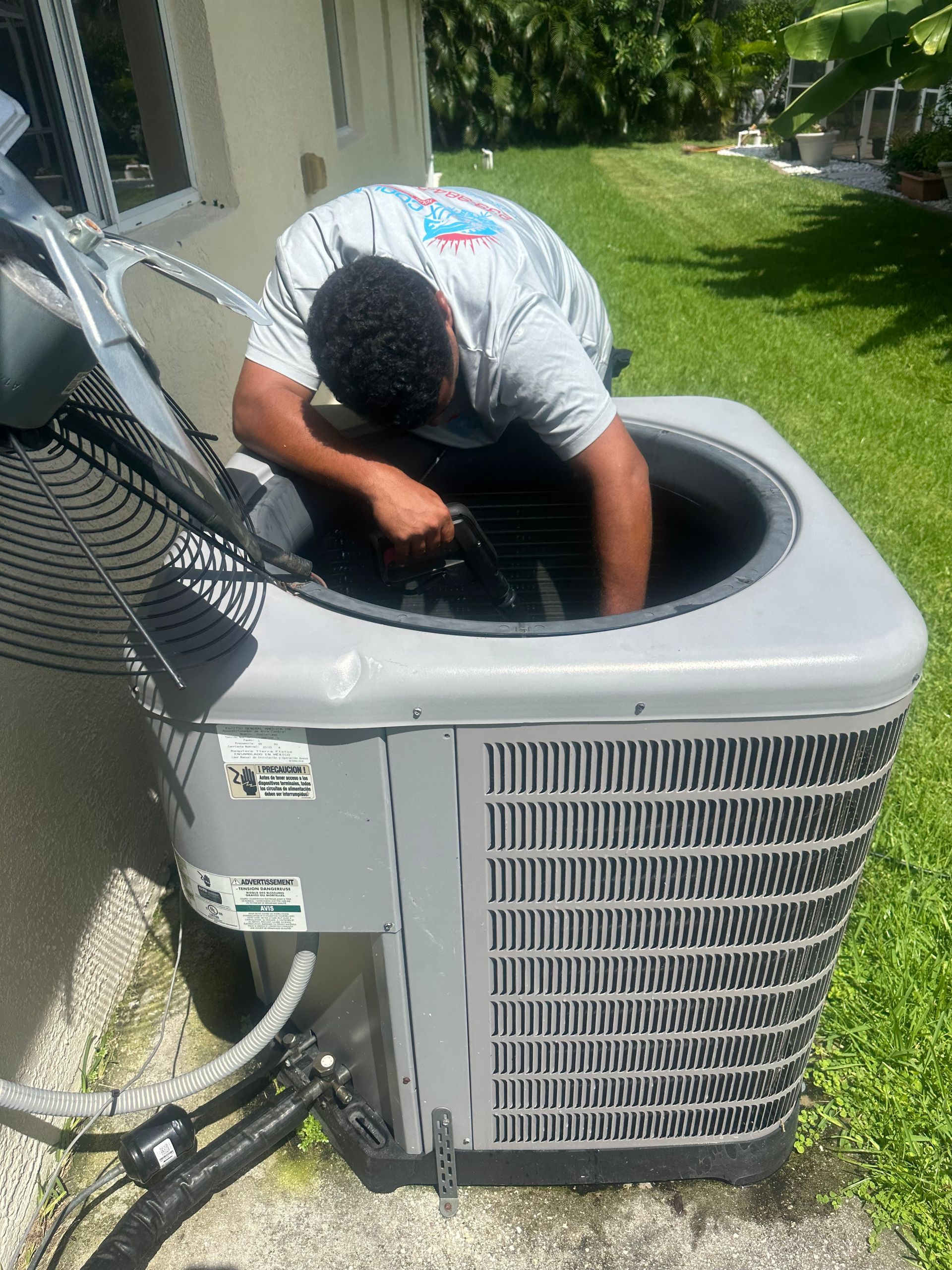 Man servicing a gray air conditioning unit outdoors. He is wearing a gray shirt, inspecting the unit. Green grass surrounds.