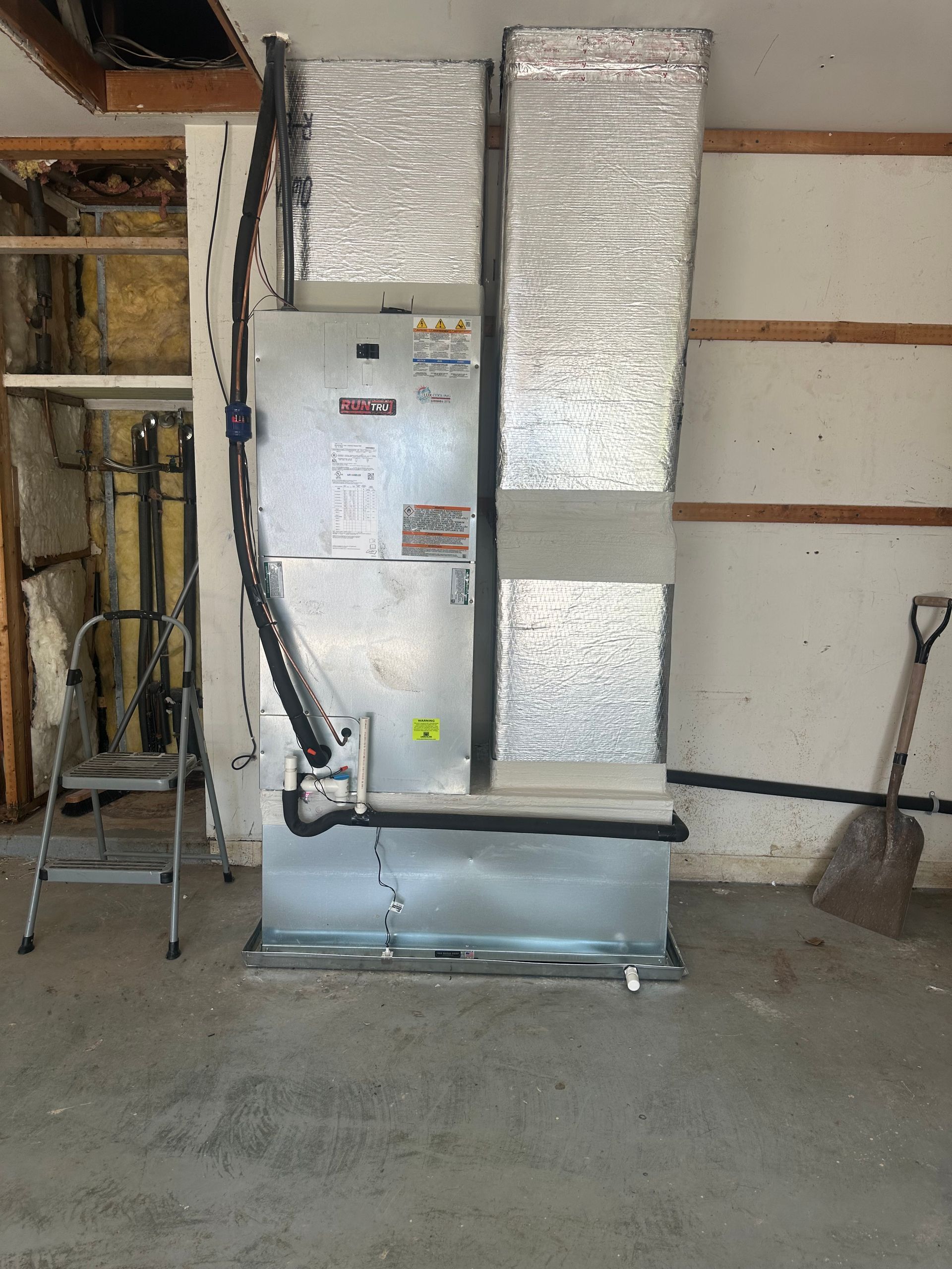 HVAC unit in a garage. Ductwork is insulated. A shovel leans against a wall.