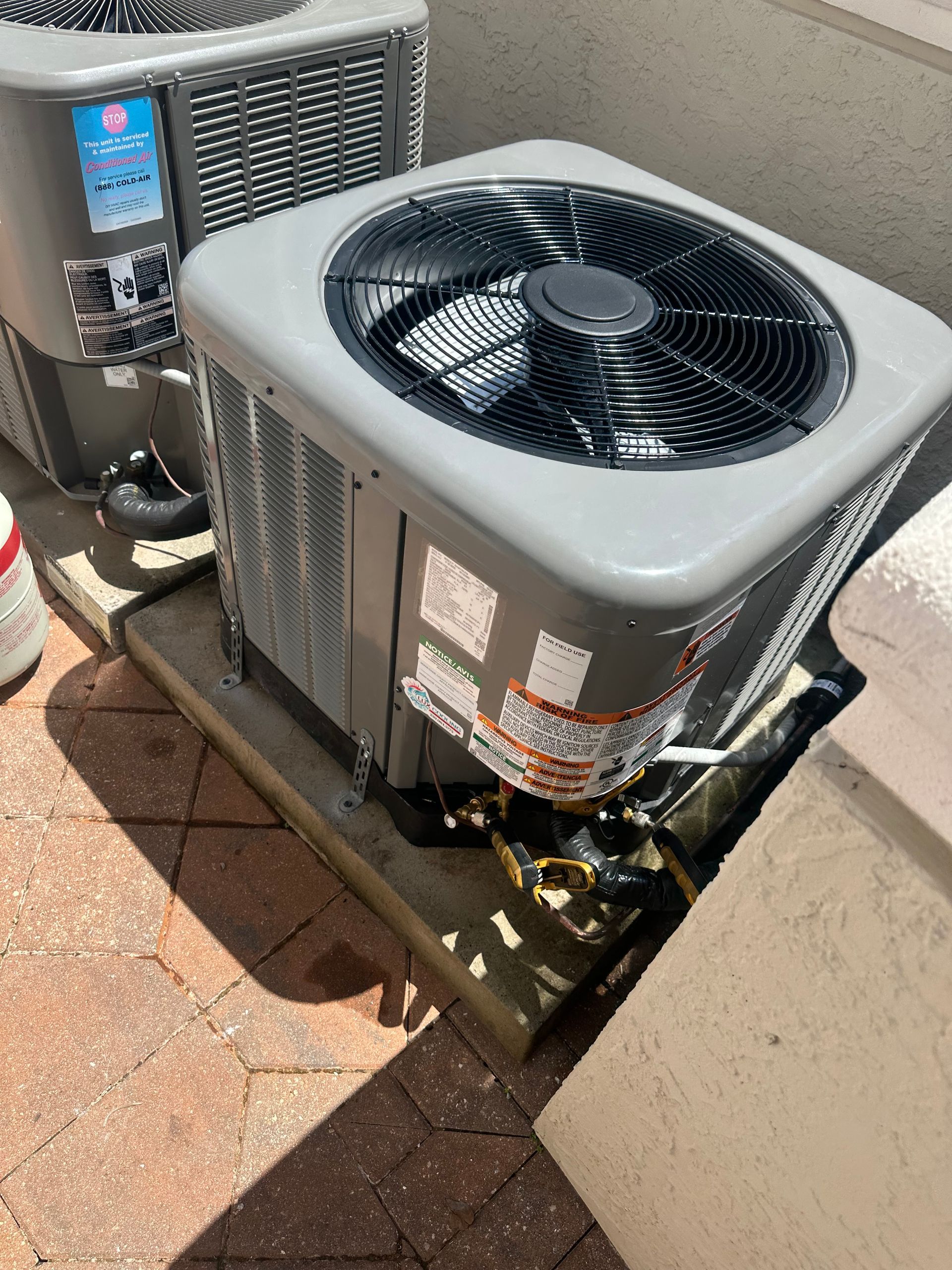 Two gray air conditioning units on a brick patio. One unit is closer and partially obscured by a concrete block.