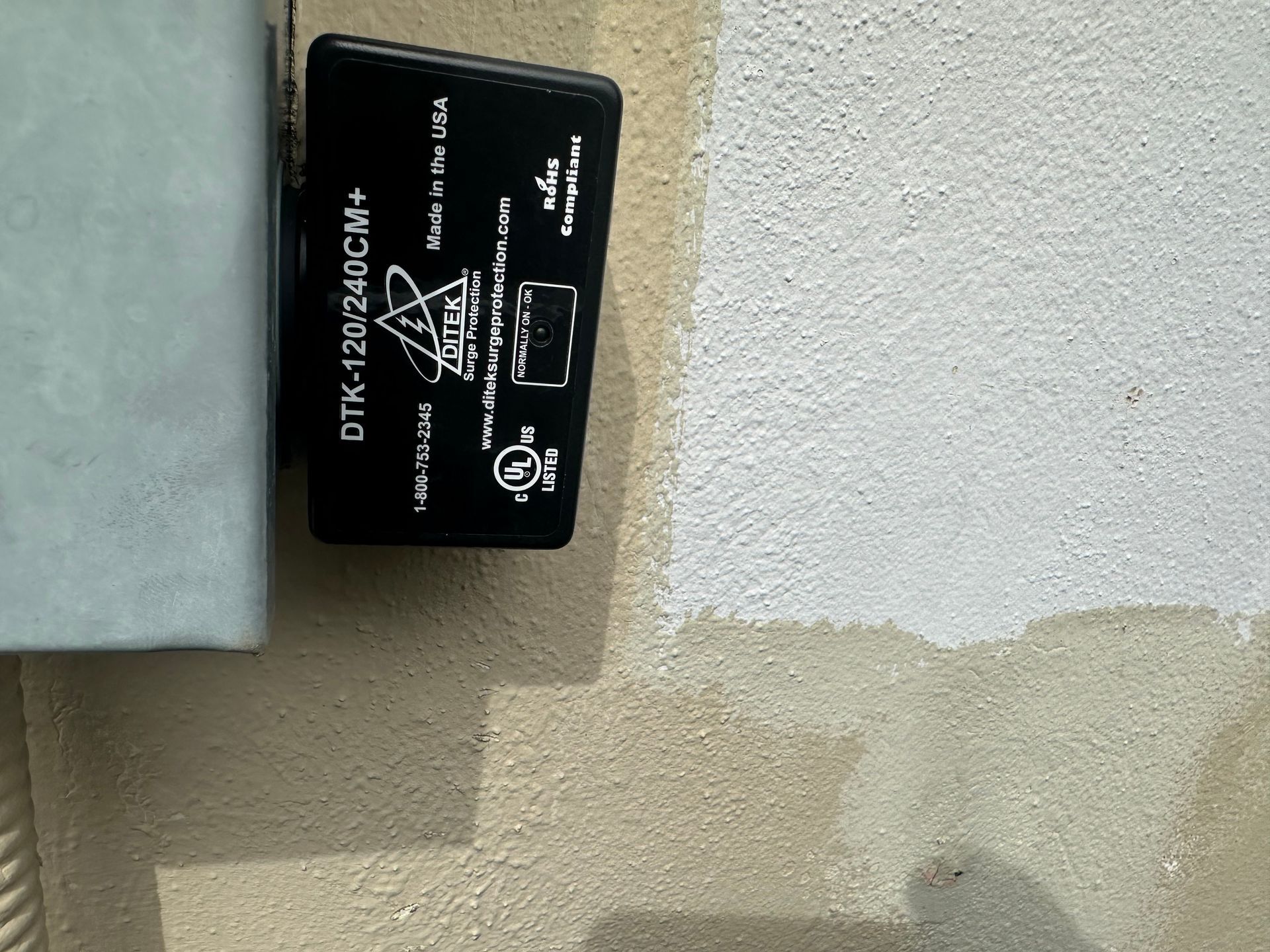 Black electrical surge protector box mounted on a white and beige stucco wall.