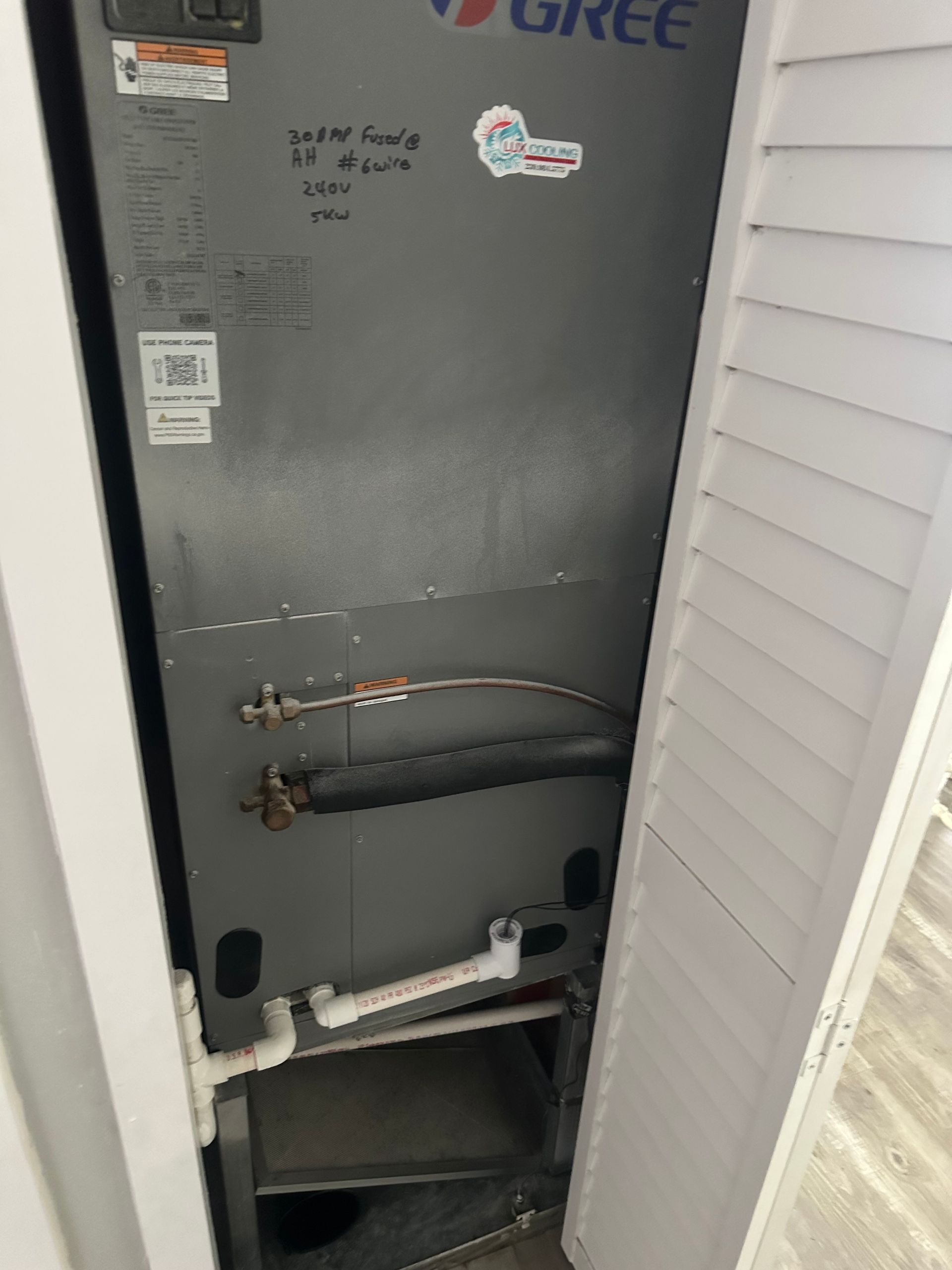A gray Gree HVAC unit in a closet, with white pipes and a partly open white louvered door.