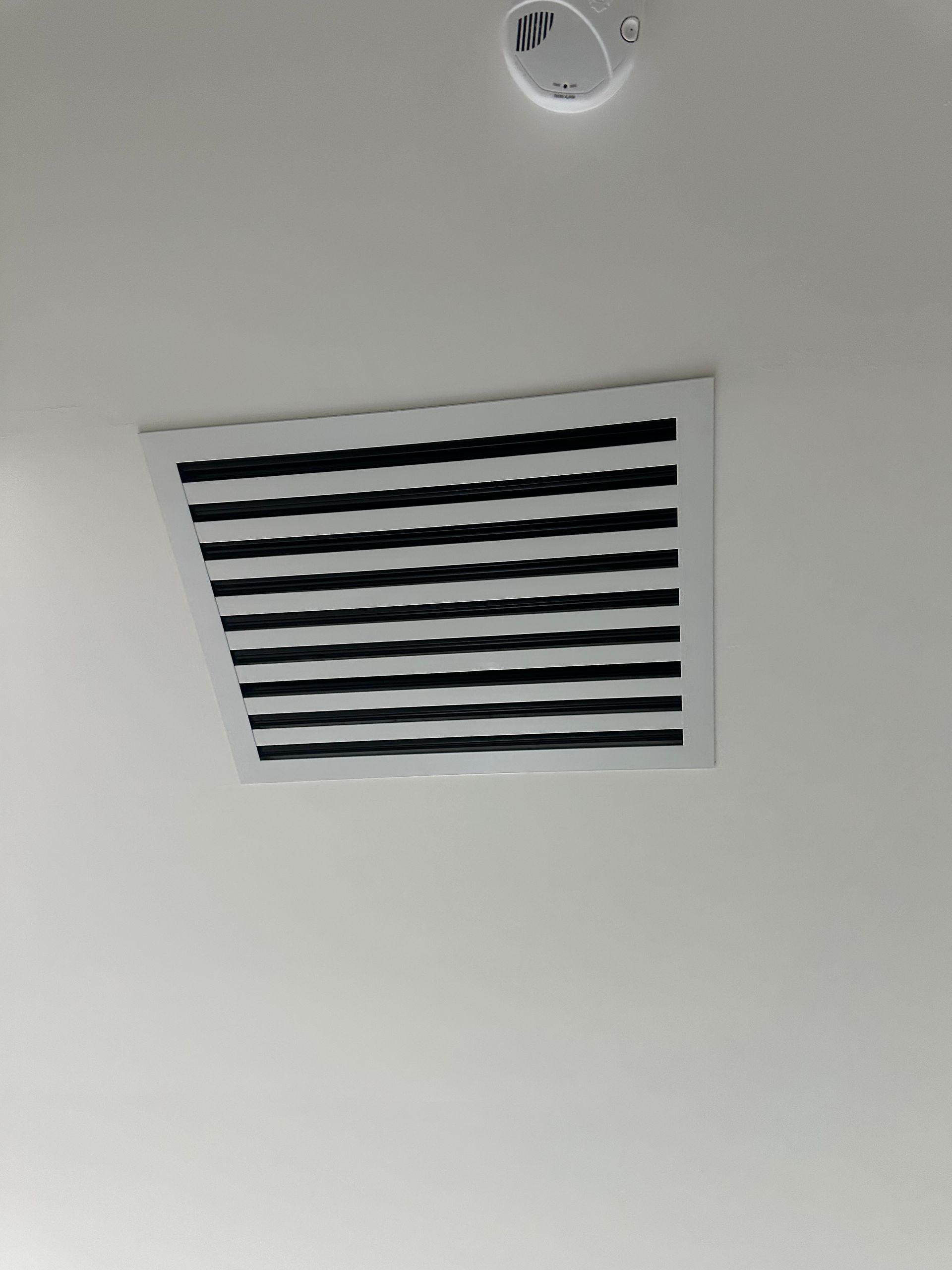 Ceiling with air vent and smoke detector. White background, black vent slats.