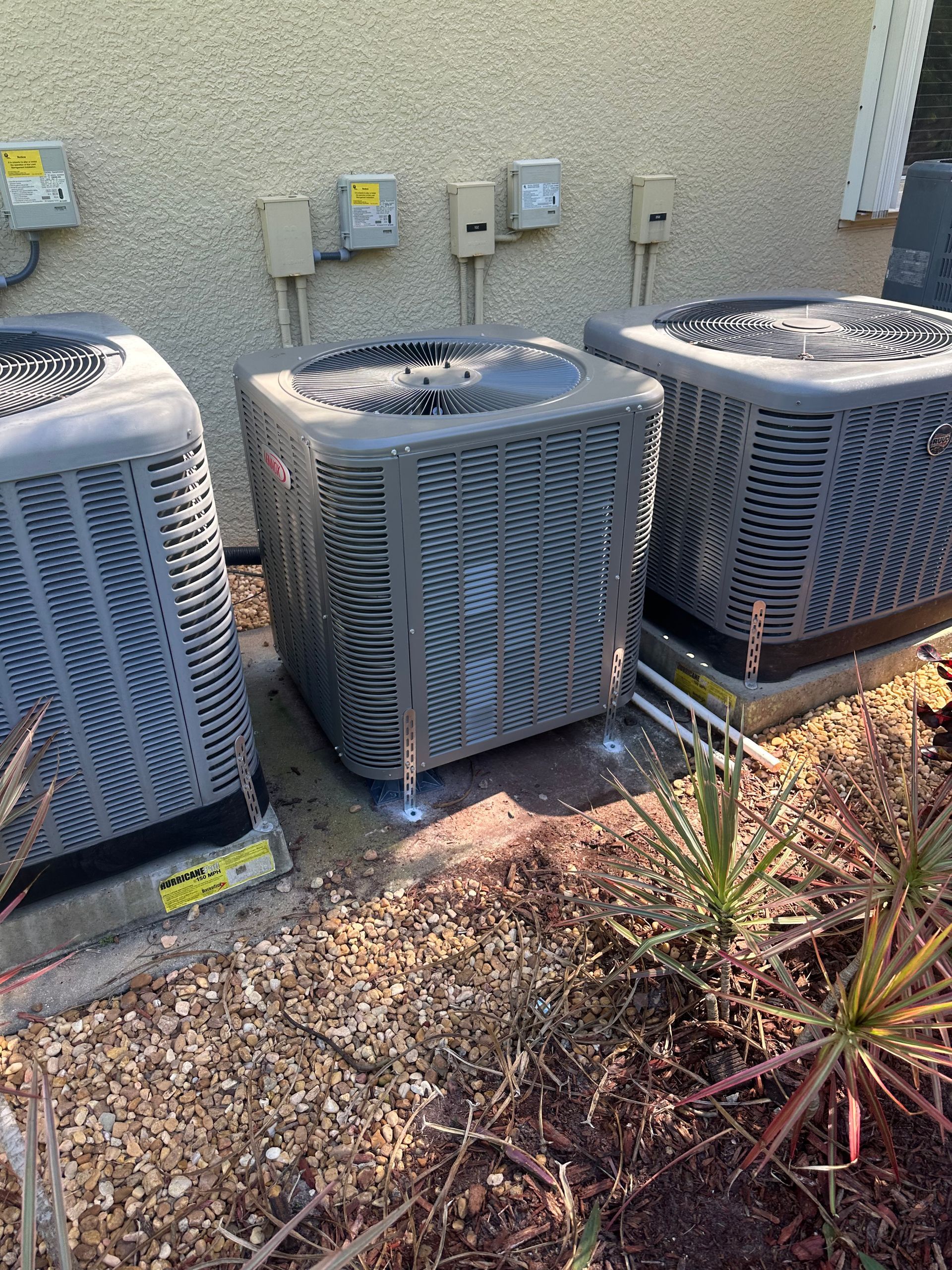 Three gray air conditioning units outside, next to a beige building with electrical boxes and some plants.