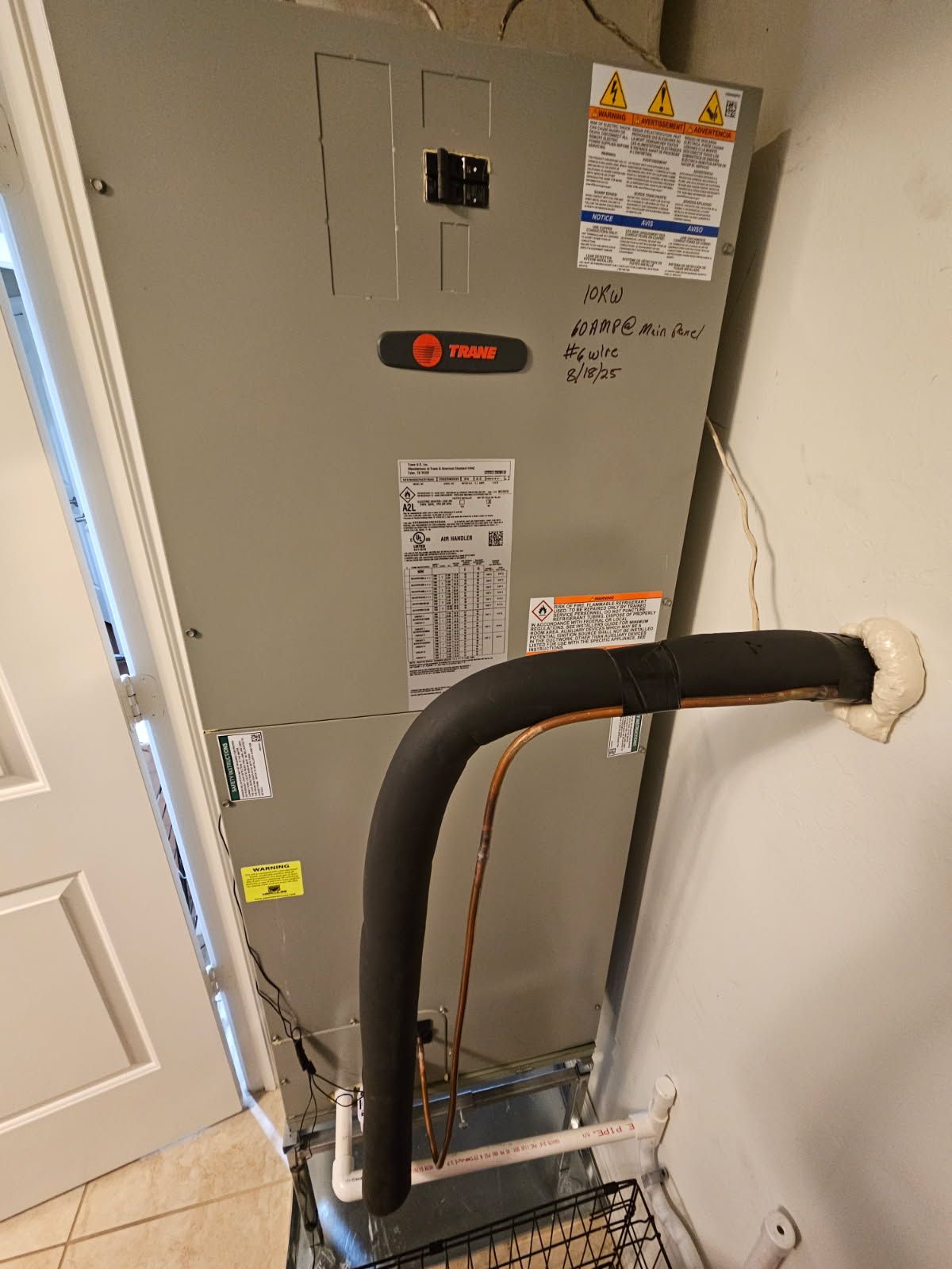 A tall, gray Trane HVAC unit, piping, and wiring against a wall with a door.
