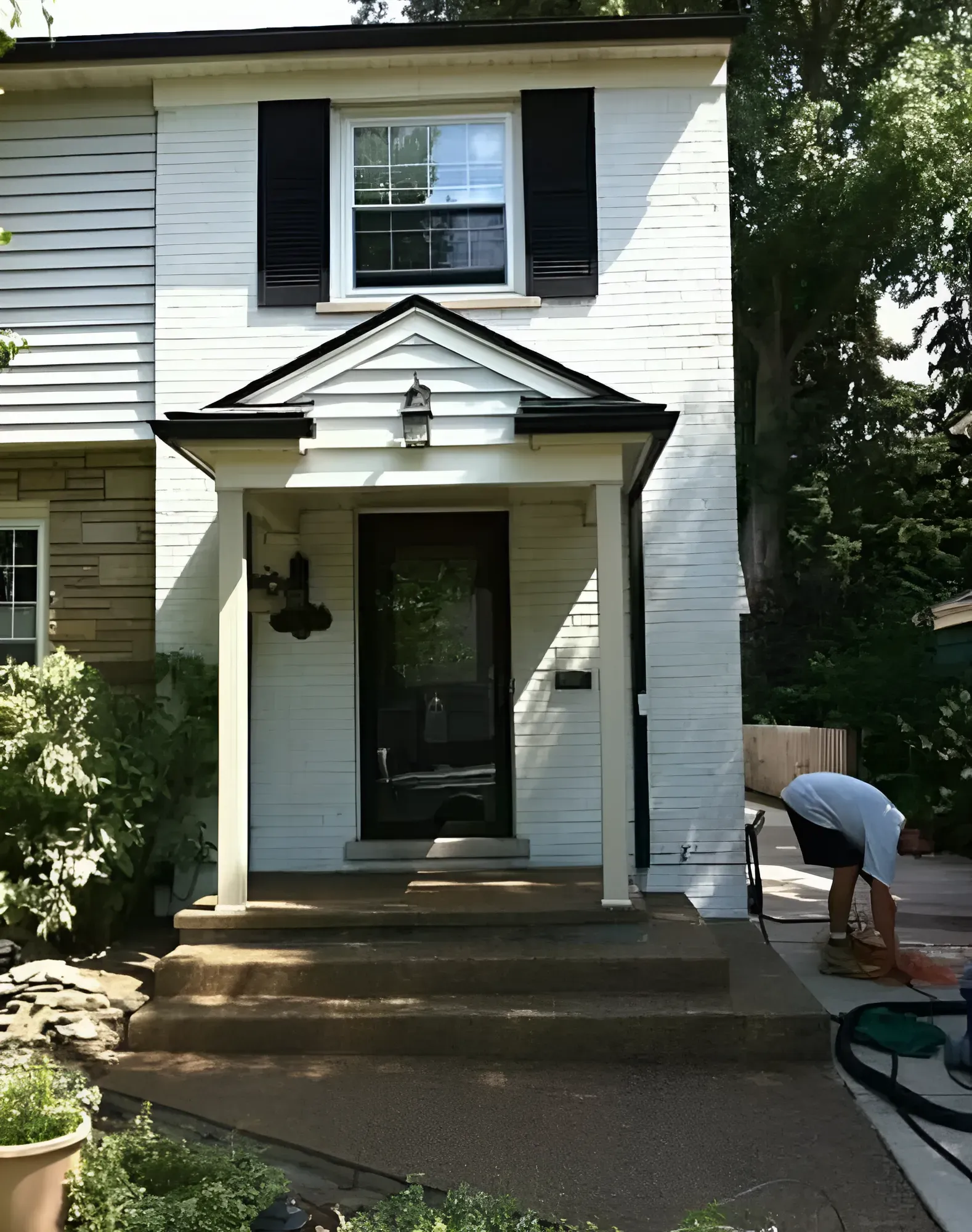 White house with black shutters and a porch, person sweeping the sidewalk.