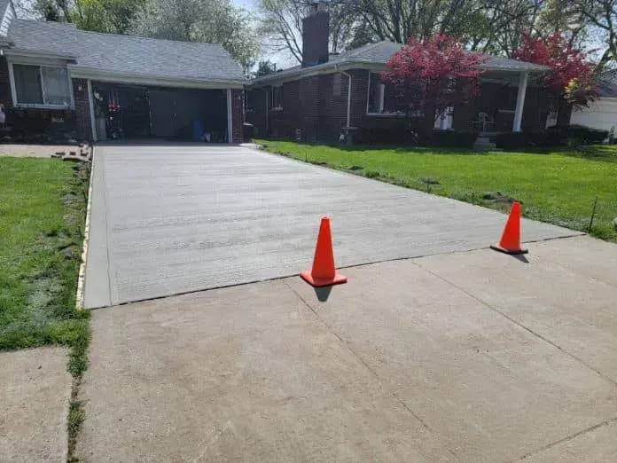 Newly poured concrete driveway with orange cones; brick house.