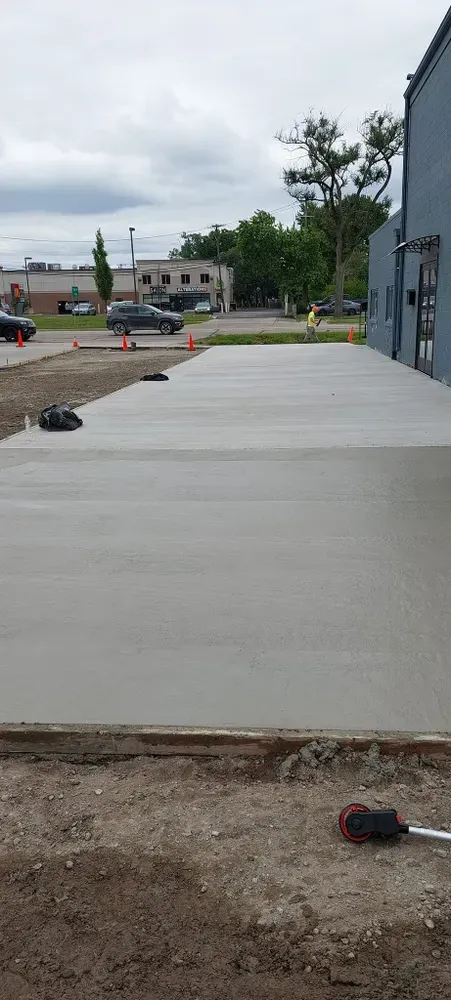 Newly poured concrete sidewalk, with construction tools and surrounding dirt, next to a building. Cars and trees in the background.
