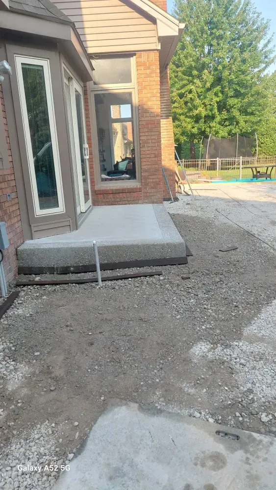 Back door with concrete patio and gravel pathway; brick building and trees in the background.