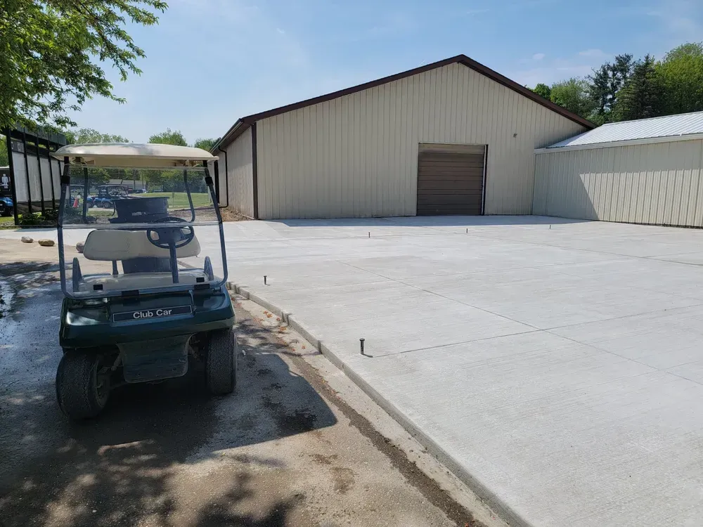 Green golf cart on a new concrete pad in front of a large tan building with a brown door. Sunny day.