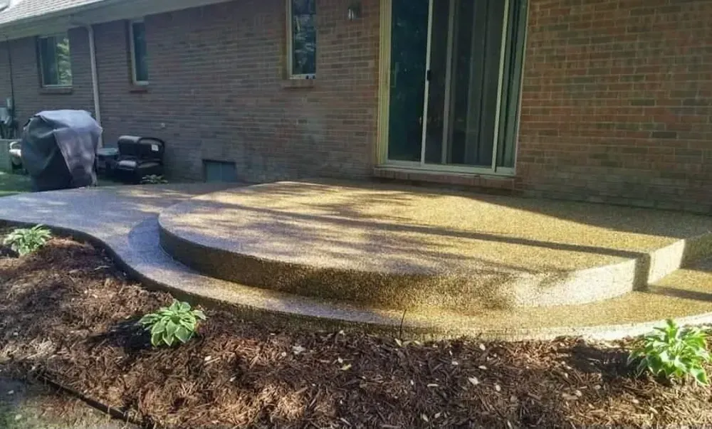 Curved concrete patio with steps, leading to a sliding glass door on a brick house, with mulch and plants.