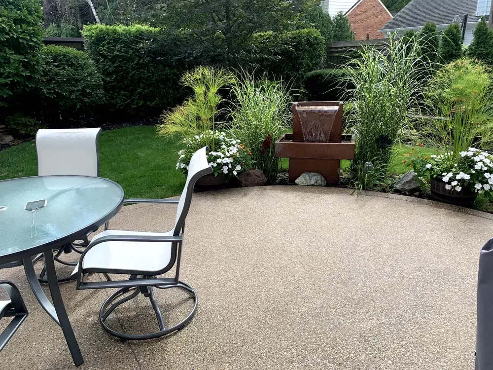 Patio with round glass table, chairs, and a small water fountain surrounded by plants and greenery.