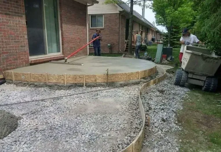 Construction of a circular concrete patio; workers smoothing, gravel base, brick house in background.