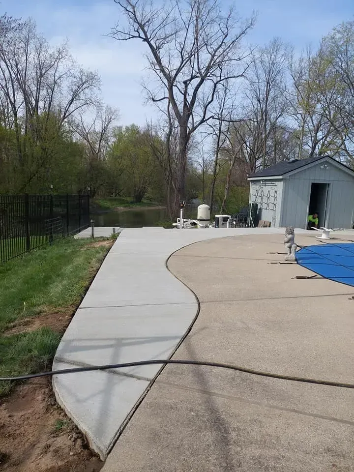 Concrete walkway curves near a pool and a body of water, with a shed in the background.