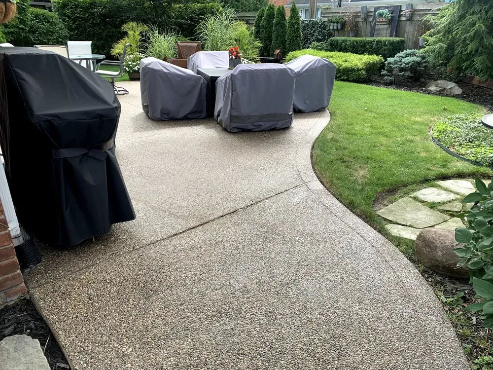 Patio with covered furniture, a grill, and a curved edge to the lawn in a backyard.